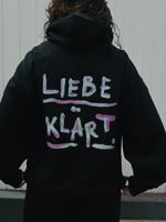 Lade das Bild in den Galerie-Viewer, Schwarzer Hoodie mit dem Schriftzug Liebe Klärt. in hell türkis und lila.
